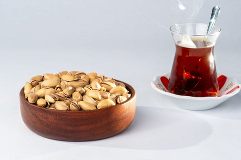 Wooden bowl of shelled pistachios next to a glass of tea on a white background